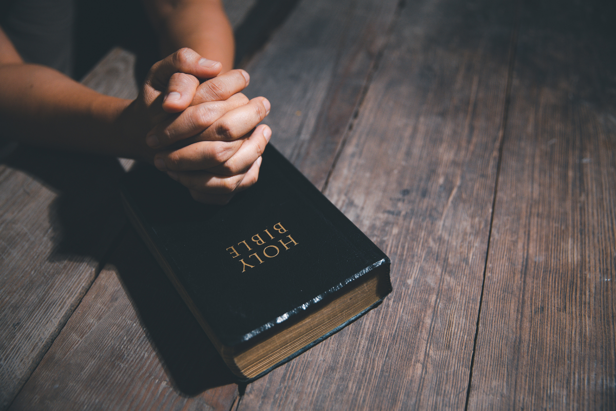 Hands clasped in prayer resting on a Bible placed on a wooden table, symbolizing faith and reflection.