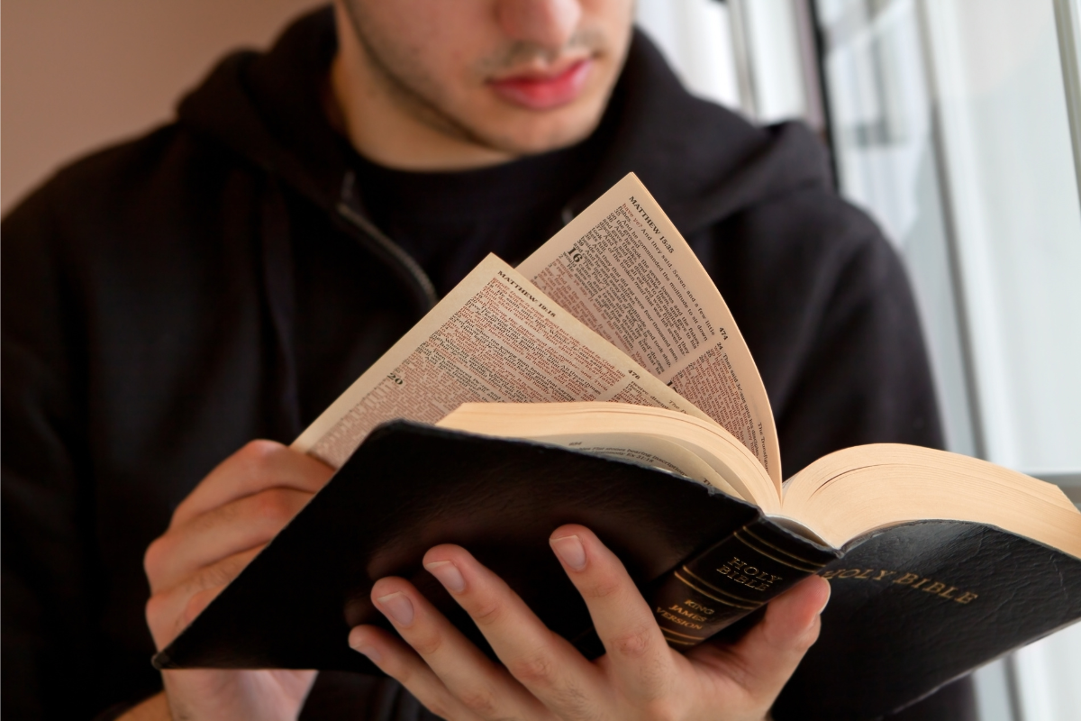 A person in a hoodie reads and closely examines a Bible, holding it open with both hands.