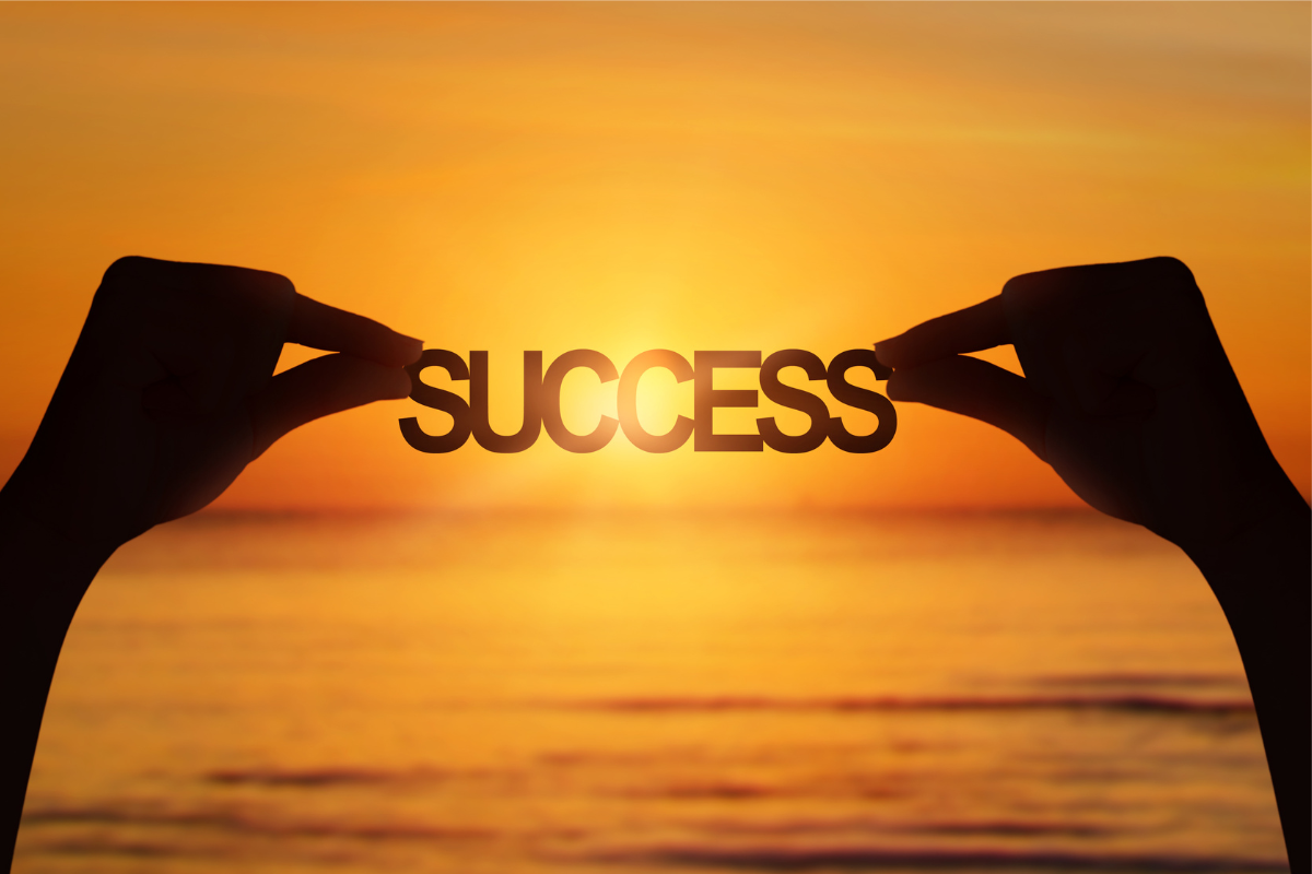 An image of a hand holding a paper-cut of the word Success over the sunset.