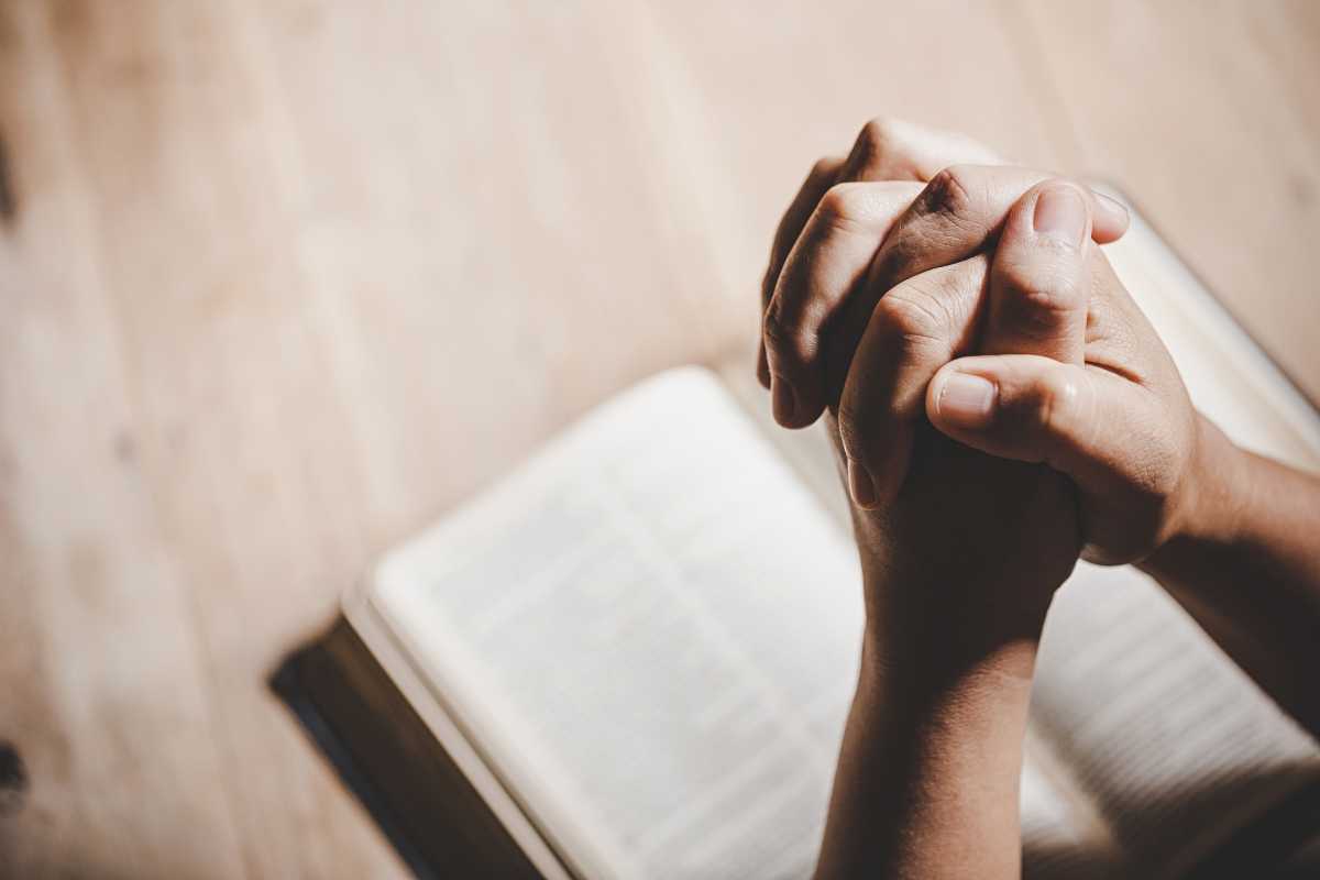 An image of hands praying with a Bible in the background.